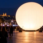 Gran esfera publicitaria inflable e iluminada internamente que brilla en una plaza al aire libre durante un evento nocturno con siluetas de la multitud