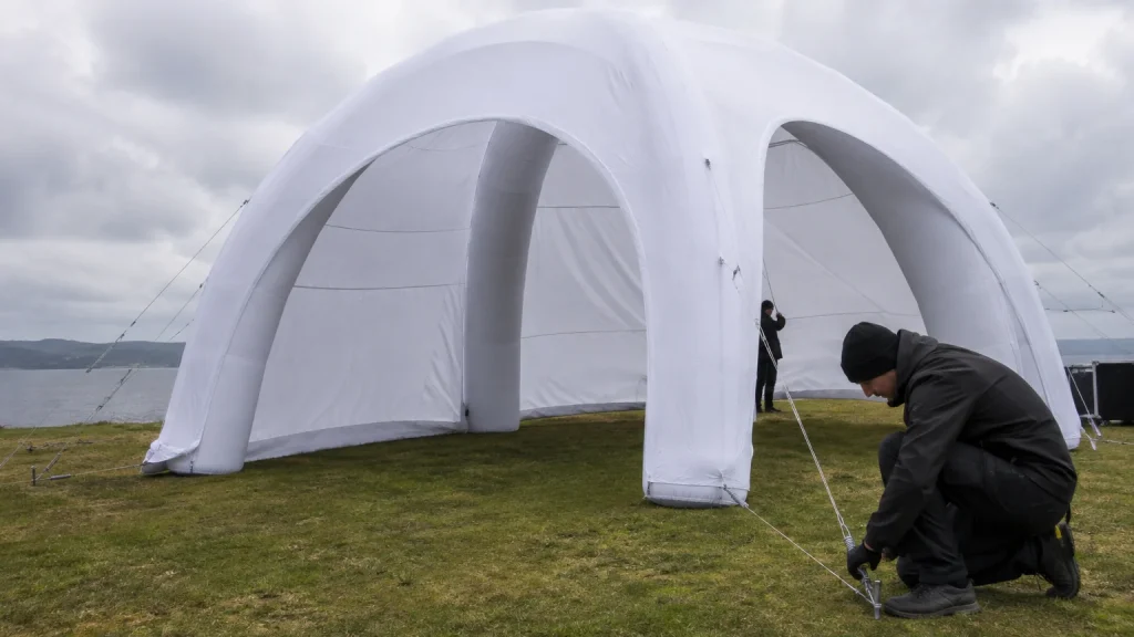Two workers anchoring a white inflatable dome tent with guy lines and ground stakes on grass by water