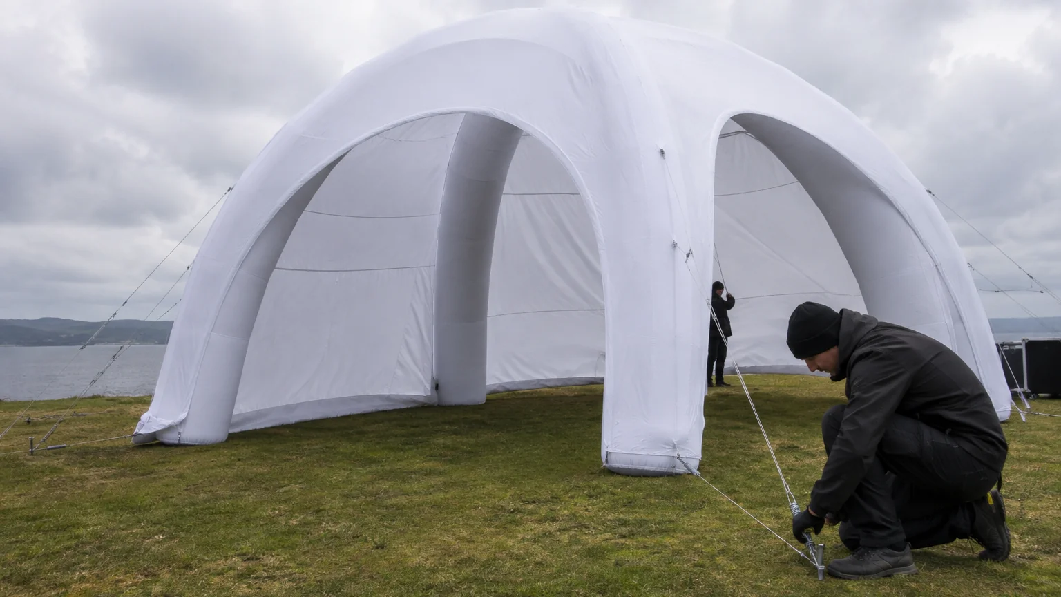Two workers anchoring a white inflatable dome tent with guy lines and ground stakes on grass by water